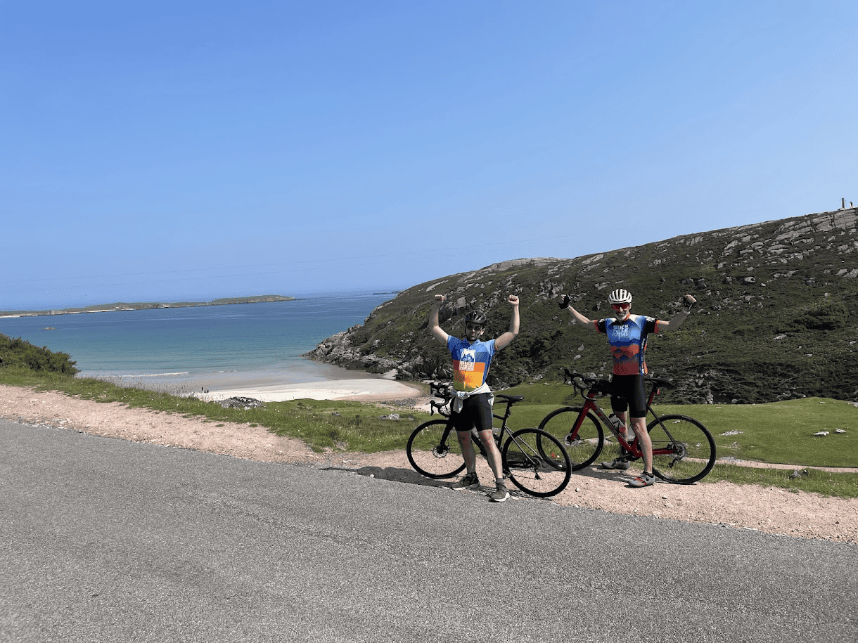 Two cyclists in Bike the UK for MS jerseys celebrating beside their bikes on a coastal road, with a sandy beach, turquoise sea and rocky hills behind them.