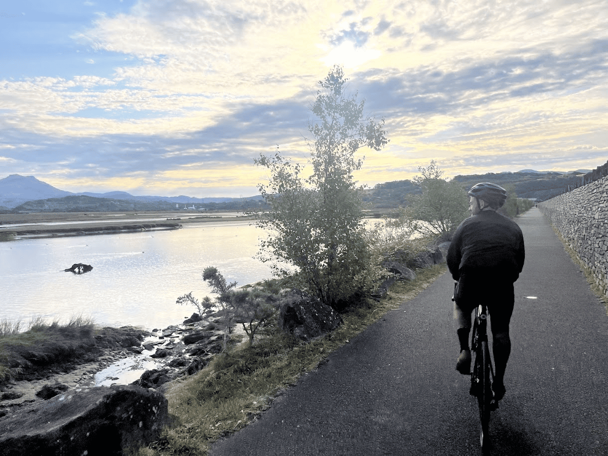 Cyclist riding along a paved waterside path beside a stone wall, with calm water, distant mountains and a cloudy sunset sky.