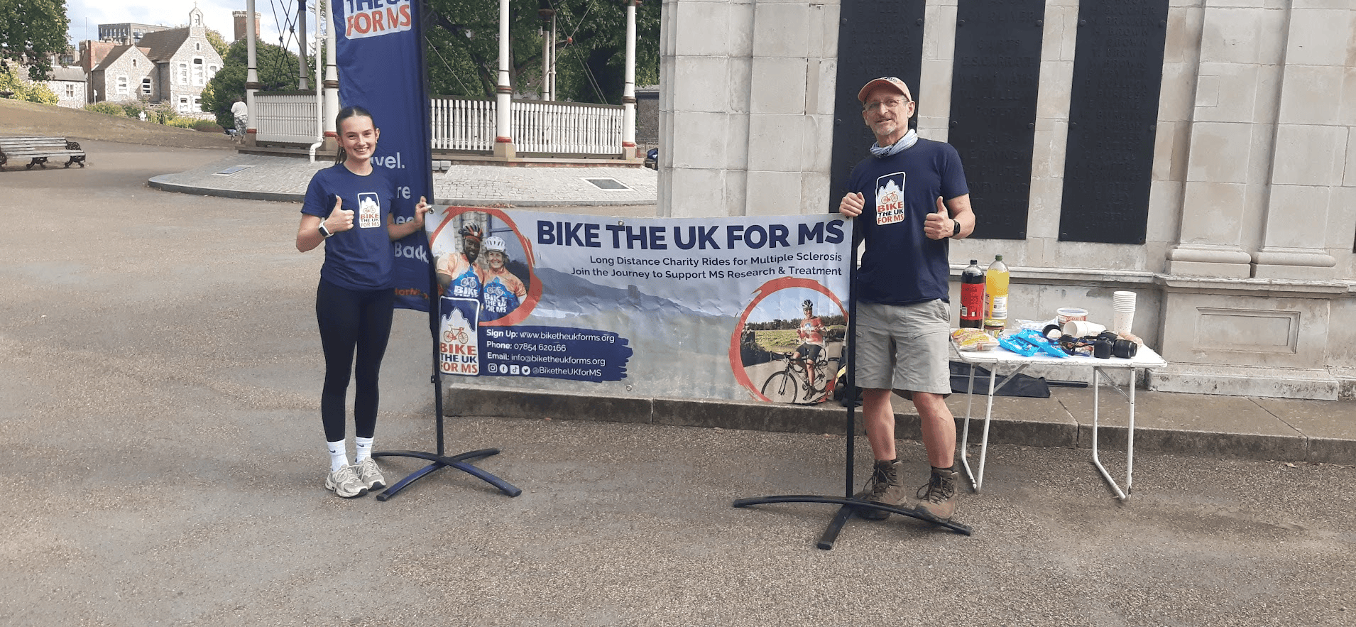 Two Bike the UK for MS volunteers giving thumbs up beside a charity banner and refreshment table at an outdoor event.