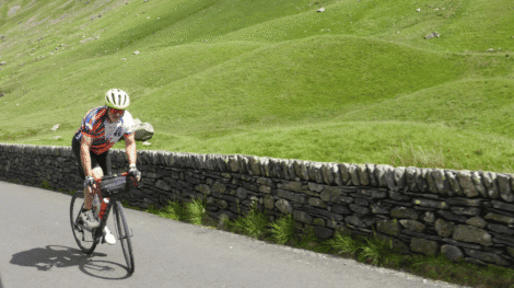Cyclist wearing a Bike the UK for MS jersey riding uphill on a country road beside a dry-stone wall, surrounded by rolling green hills.