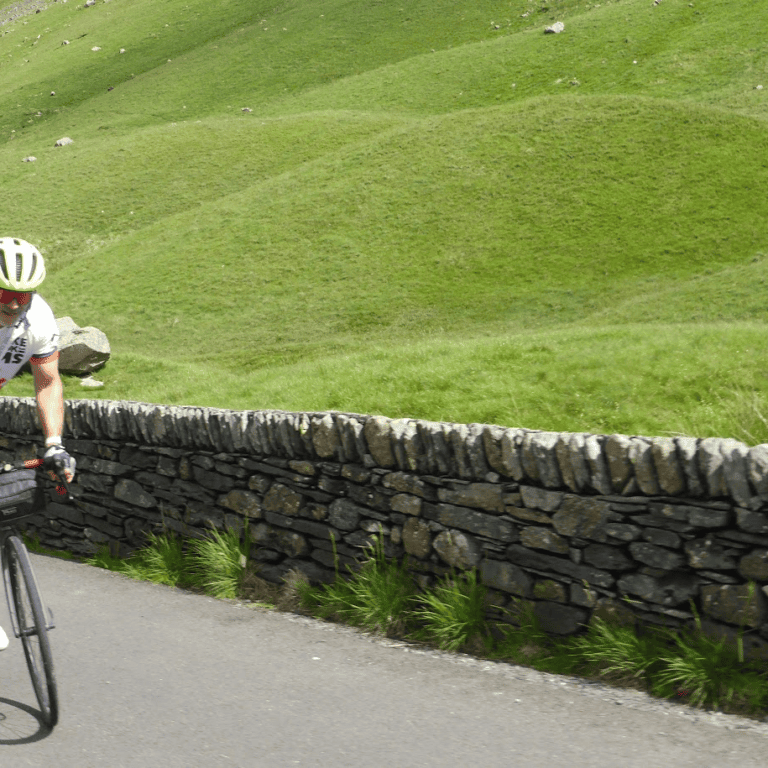 Cyclist wearing a Bike the UK for MS jersey riding uphill on a country road beside a dry-stone wall, surrounded by rolling green hills.