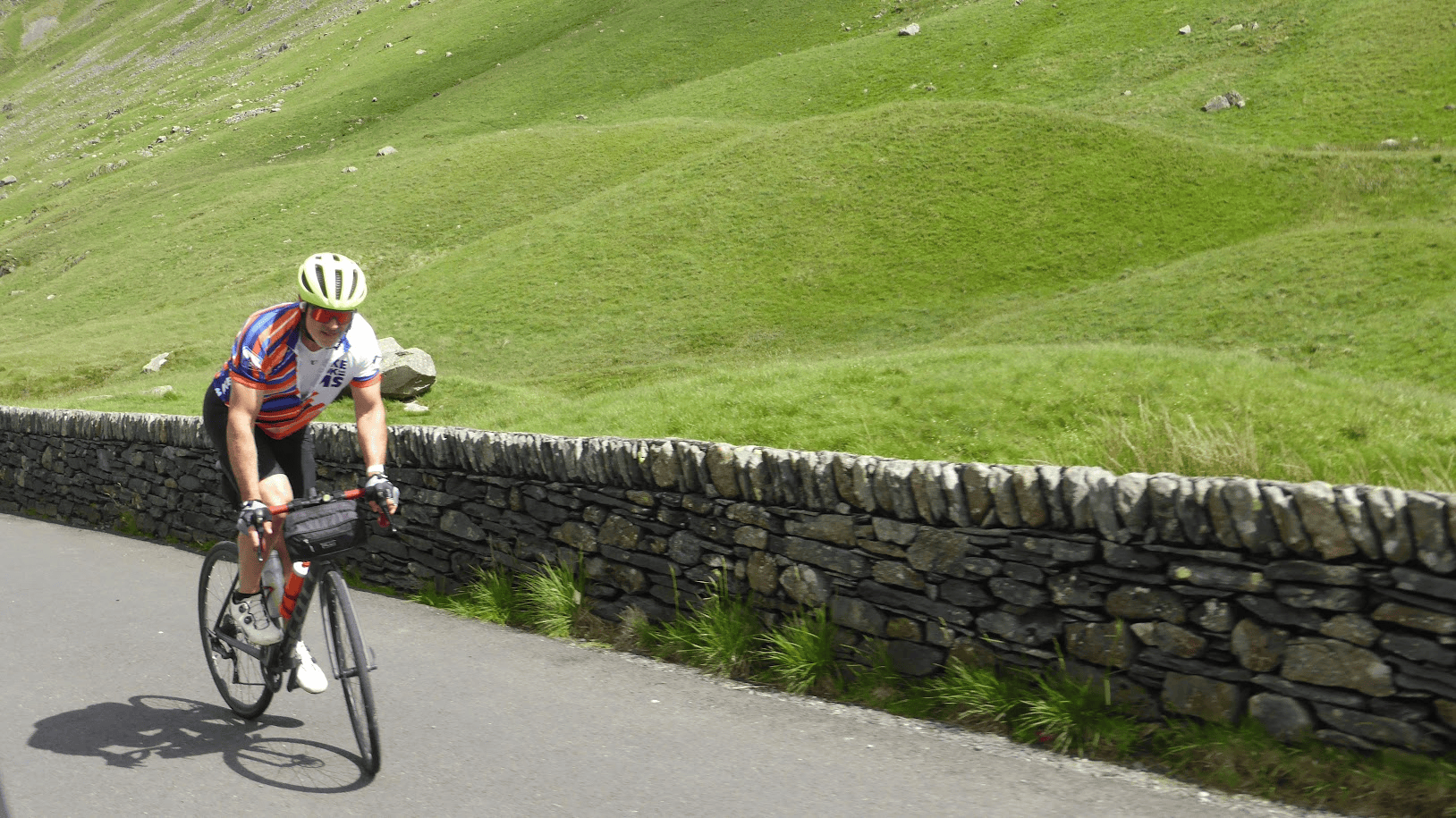 Cyclist wearing a Bike the UK for MS jersey riding uphill on a country road beside a dry-stone wall, surrounded by rolling green hills.