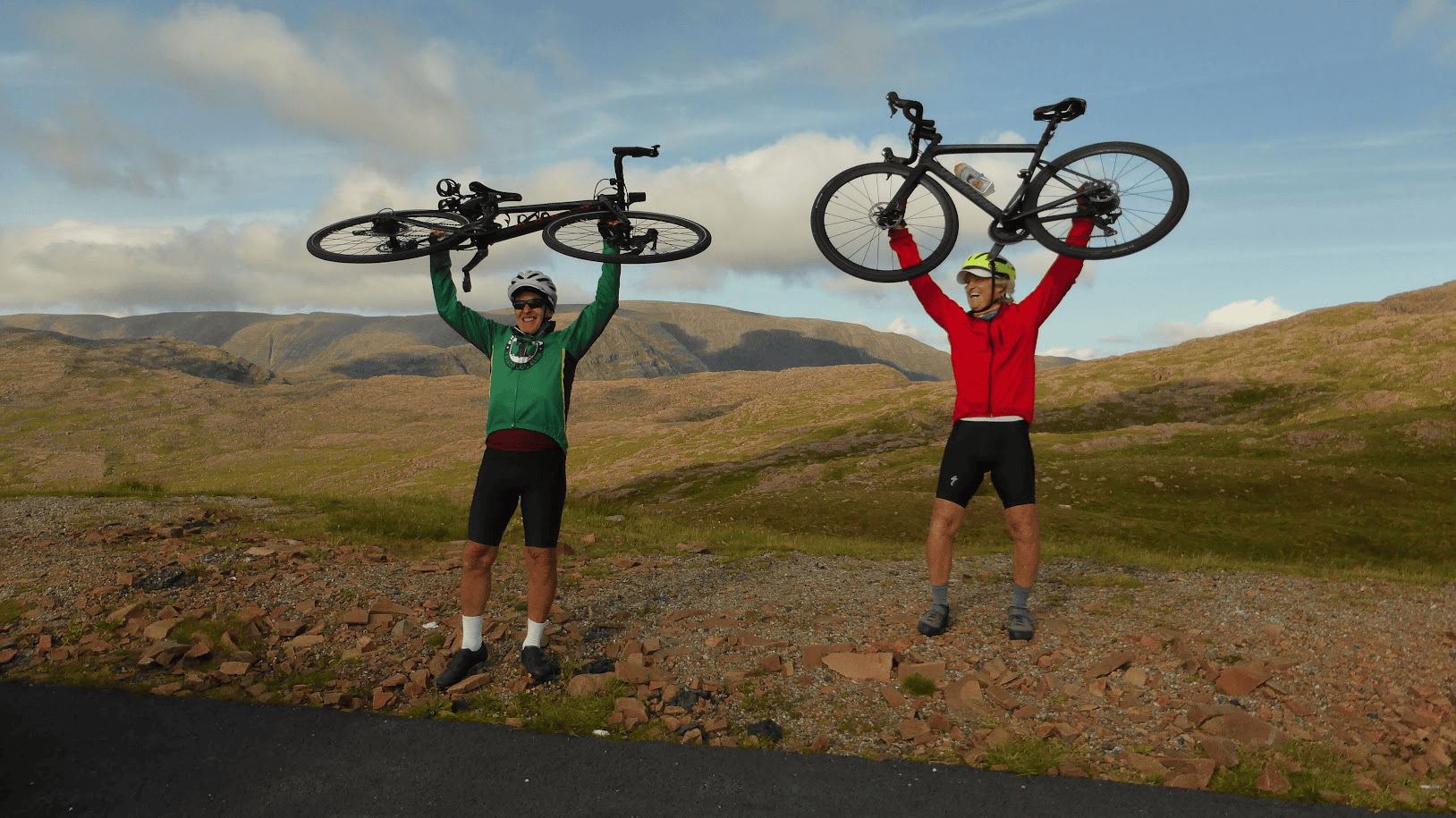 Two cyclists celebrating with their bikes held overhead against a backdrop of rugged mountain scenery.