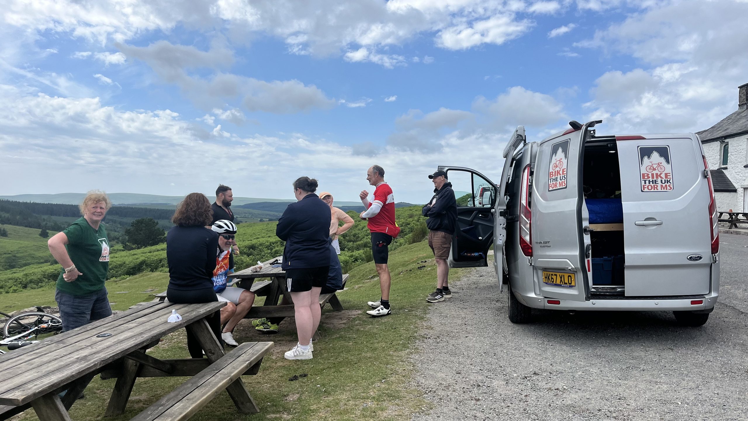 Bike the UK for MS van parked for a rest stop with cyclists and route leaders standing around and beautiful views of the countryside behind them